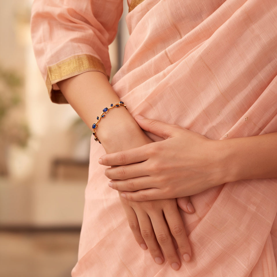 A woman's wrist in a peach saree sleeve with a gold border, wearing the blue tulip gold tone bracelet