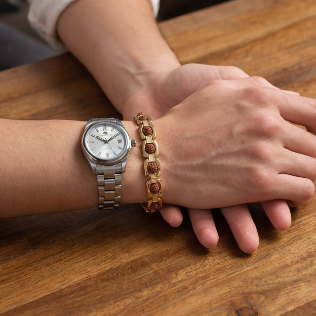 Man's wrist wearing a gold plated rudraksha bracelet beside a steel wristwatch