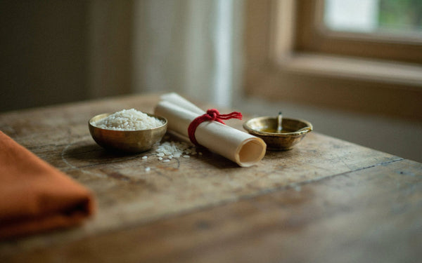 A rolled paper scroll tied with red thread beside a brass diya and a small brass bowl of rice on a wooden table