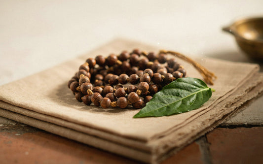 Tulsi wood mala with small brown beads coiled on a sand cloth beside a tulsi leaf