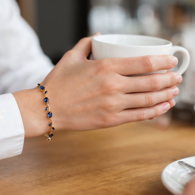 A woman's hand around a coffee cup at a cafe table, wearing the blue tulip gold tone bracelet