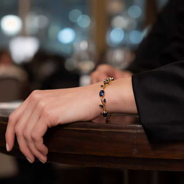 A woman's hand and wrist in a black satin sleeve resting on a dark wooden table at an evening event, wearing the blue tulip gold tone bracelet