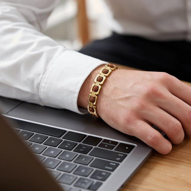 Man's wrist with a gold link Rudraksha bracelet beside a laptop at a wooden desk